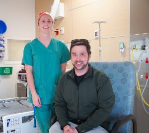 Almir is a burns patient at the royal Adelaide hospital who is sitting on a chair in his private hospital room with his nursing manager named Mikaela who is standing just behind him with her green scrubs on