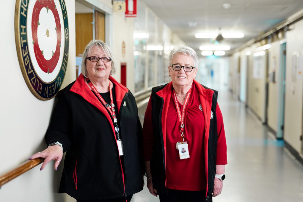 Two women who are volunteers with the Friends of The Queen Elizabeth Hospital stand in hospital corridor
