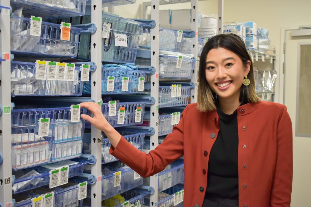 Doctor standing in pharmacy area of hospital with drugs on shelves behind her.
